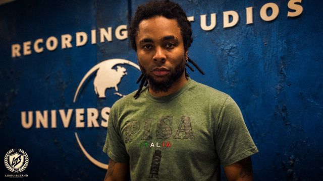 Luigui Bleand in a green T-shirt in front of a blue recording-studio wall sign, with his logo in the corner