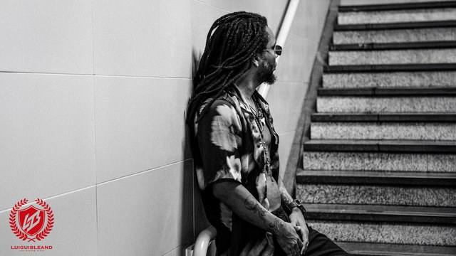 Luigui Bleand beside a staircase, in a black-and-white press photograph with his red logo